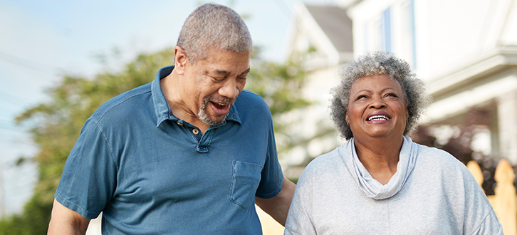 A man and woman smile and walk outdoors, side by side, on a sunny day.